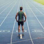 Rear view of a male sprinter standing at the starting blocks on a blue running track, representing athletic performance and health.