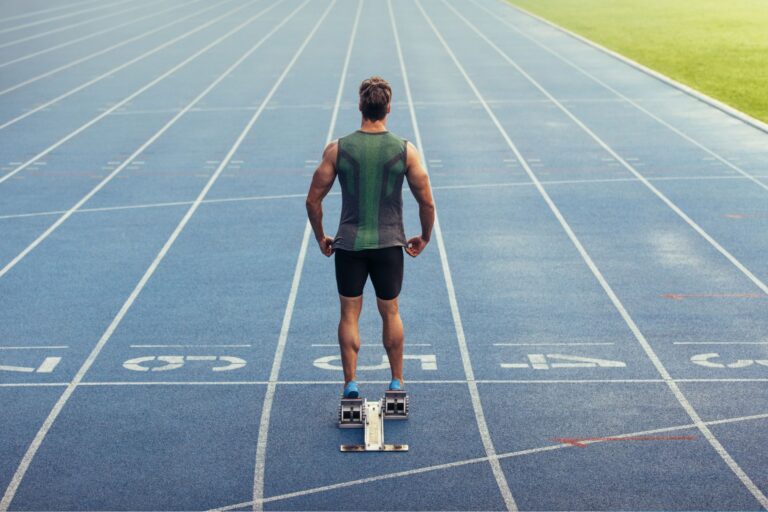 Rear view of a male sprinter standing at the starting blocks on a blue running track, representing athletic performance and health.